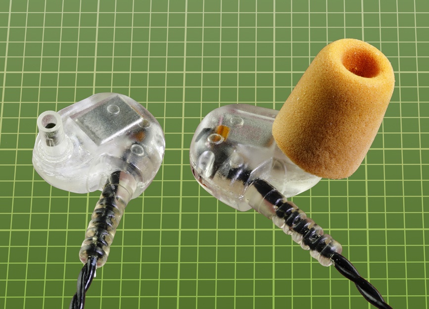 A pair of in-ear headphones, supplied with disposable foam plugs that conform flexibly with the ear canal. The headphone on the left is shown with its plug removed.