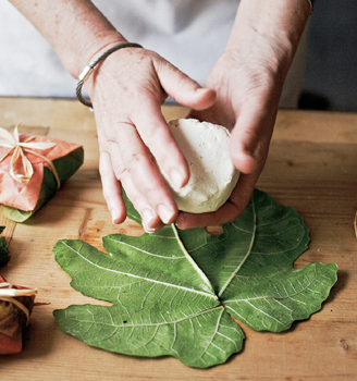 Two hands about to place a round of goat cheee on a flattened grape leaf sitting upon a wooden board