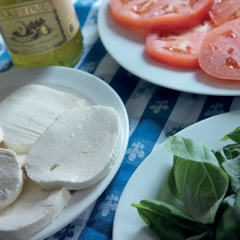 A plate of sliced tomatoes, a dish of basil leaves, and a plate of bright white fresh mozzarella cheese