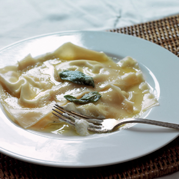 A deep white plate filled with ravioli and topped with two large sage leaves. A silver fork dripping with ricotta sits on the side of the dish.