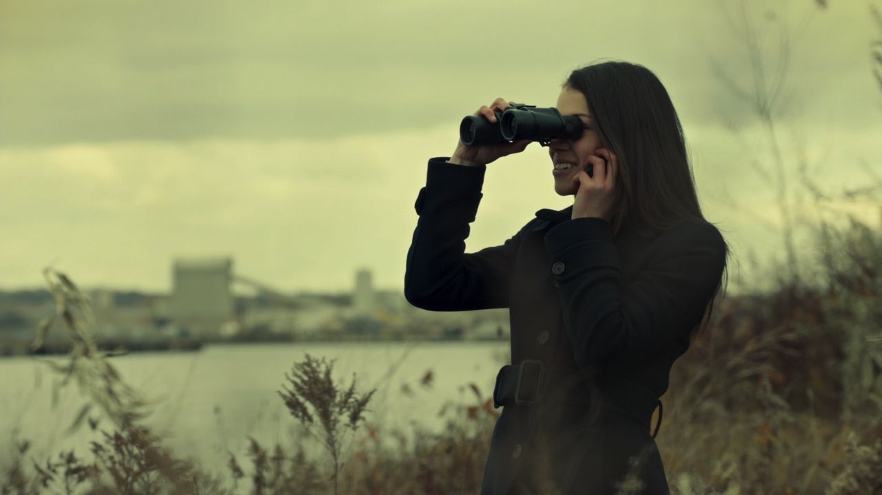 Frame from Orphan Black pilot with Sarah Manning silhouetted beside a river watching her own memorial service through binoculars.