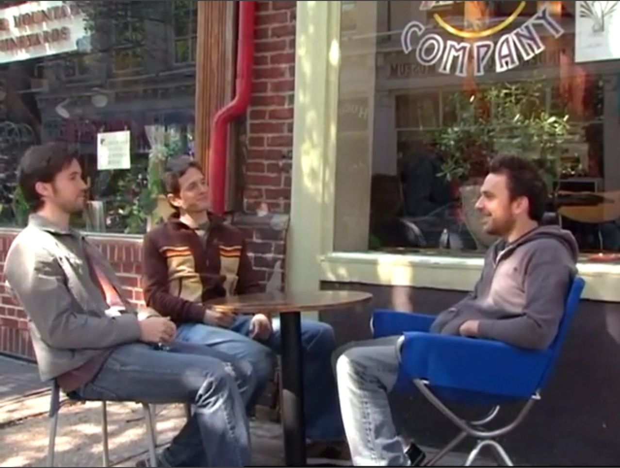 In frame from It’s Always Sunny pilot, Mac, Dennis, and Charlie sit at a sidewalk table outside a coffee shop.