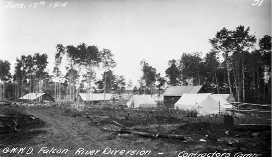 Black and white photograph of a small cluster of white tents and buildings surrounded by trees, and a dirt road in the foreground. White script at upper left: June 17th, 1914; at bottom: G.W.W.D. Falcon River Diversion – Contractors [sic] Camp.