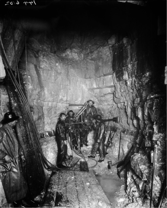 Black and white vertically oriented photograph of a group of five miners standing within a tunnel and the surrounding rough rock face; the image appears illuminated from the centre by a spotlight fading to black above, and everything, from the miners clothing to the rock face looks glistening and wet. At the far left foreground a sixth miner stands looking towards the rest beside a long cluster of black cables that runs down from the darkness above him along the rock and over towards the centre area where the other miners are grouped together. Wooden planks line the ground beside pooling water and various tools lean against the rock at the right hand side of the image.