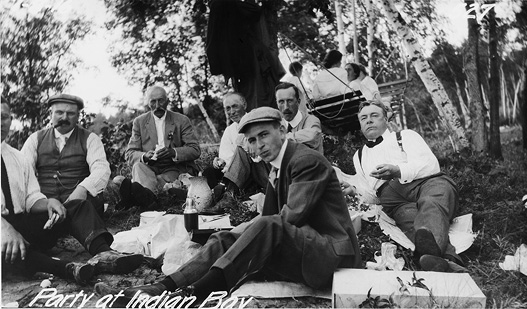 Black and white photograph of a group of seven white men picnicking outdoors. All are seated in lounging and relaxed poses on the ground and all face the camera. Three or four more people are seen sitting on a hammock behind them near surrounding trees, facing away from the camera. White script at bottom reads: Party at Indian Bay.