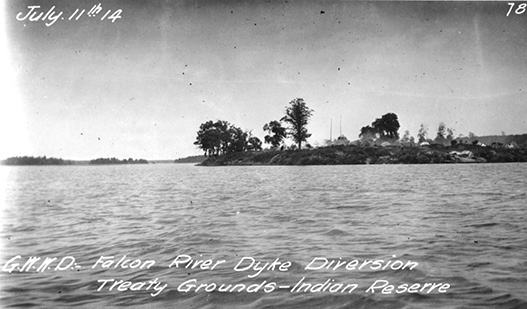 Black and white photograph of a land mass at centre and right with some trees and rocks surrounded by water, and more land behind it along the horizon. Water takes up the foreground, and a clouded sky rests above. White script at upper left: July 11th, 14; and at bottom: G.W.W.D. Falcon River Dyke Diversion Treaty Grounds – Indian Reserve.