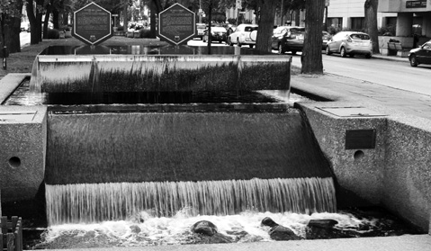 Black and white photograph of a section of a concrete monument featuring three tiers with a waterfall flowing down along each wide concrete tier, and two hexagonal plaques affixed to the top tier. From the tree trunks and flanking streets beyond one can discern that the monument is located at the centre of a boulevard. Water flows over large rocks at its lower echelon.