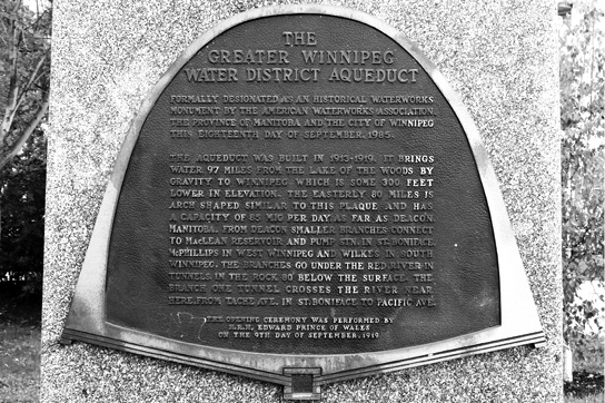 A black and white photograph of a metal plaque affixed to stone, in the shape of the aqueduct’s arch. Upon the plaque is the following text in relief: THE GREATER WINNIPEG WATER DISTRICT AQUEDUCT / FORMALLY DESIGNATED AS AN HISTORICAL WATERWORKS MONUMENT BY THE AMERICAN WATERWORKS ASSOCIATION, THE PROVINCE OF MANITOBA AND THE CITY OF WINNIPEG THIS EIGHTEENTH DAY OF SEPTEMBER, 1985. THE AQUEDUCT WAS BUILT IN 1913-1919. IT BRINGS WATER 97 MILES FROM THE LAKE OF THE WOODS BY GRAVITY TO WINNIPEG WHICH IS SOME 300 FEET LOWER IN ELEVATION. THE EASTERLY 80 MILES IS ARCH SHAPED SIMILAR TO THIS PLAQUE AND HAS A CAPACITY OF 85 M10 PER DAY AS FAR AS DEACON, MANITOBA. FROM DEACON SMALLER BRANCHES CONNECT TO MACLEAN RESERVOIR AND PUMP STN. IN ST. BONIFACE, MCPHILLIPS IN WEST WINNIPEG AND WILKES IN SOUTH WINNIPEG. THE BRANCHES GO UNDER THE RED RIVER IN TUNELS IN THE ROCK 80 BELOW THE SURFACE. THE BRANCH ONE TUNNEL CROSSES THE RIVER NEAR HERE FROM TACHE AVE. IN ST. BONIFACE TO PACIFIC AVE. / THE OPENING CEREMONY WAS PERFORMED BY H.R.H. EDWARD PRINCE OF WALES ON THE 9TH DAY OF SEPTEMBER 1919.