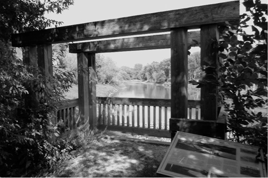 Black and white photograph taken from within a wooden decked area, with wooden railing and heavy beams standing to create parallel rectangular archways. The area overlooks a river with overgrown banks on a sunny day. Plants lean in from the sides of the frame in the foreground and in the right foreground stands a plaque.