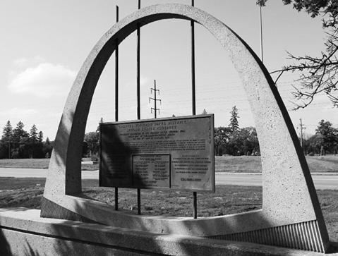 Black and white photograph on a sunny day of a sculptural monument made from concrete in the shape of a slice of aqueduct, with what looks to be a steel plaque affixed to its centre opening with three vertical-running steel rods. A road, hydro lines, and trees are visible in the background.