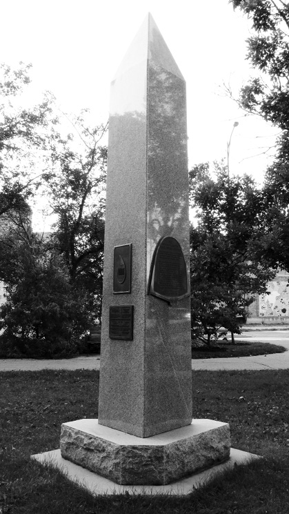 A black and white vertically-oriented photograph of what looks to be a granite monument in the shape of an obelisk. Upon one face of it is the aqueduct arch-shape, and two plaques are seen affixed to an adjacent face. Grass surrounds the wider base of the structure, with a sidewalk and trees behind.