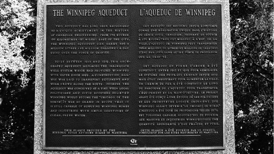 Black and white photograph of rectangular metal plaque affixed to stone. English text appears at the left and French at the right with the headings: THE WINNIPEG AQUEDUCT and L’AQUEDUC DE WINNIPEG, and the following body of text: THIS AQUEDUCT HAS LONG BEEN RECOGNIZED AS A UNIQUE ACHIEVEMENT IN THE HISTORY OF CANADIAN ENGINEERING. FROM ITS SOURCE 156 KILOMETRES (197 MILES) EAST OF THE CITY, THE WINNIPEG AQUEDUCT CAN CARRY 386.4 MILLION LITRES (85 MILLION GALLONS) A DAY USING ONLY THE FORCE OF GRAVITY. BUILT BETWEEN 1913 AND 1919, THIS ARCH-SHAPED AQUEDUCT REPLACED THE INADEQUATE WELL SYSTEM WHICH HAD PROVIDED WINNIPEG WEITH WATER SINCE 1899. A CONSTRUCTION RAILWAY WAS LAID TO TRANSPORT EQUIPMENT AND WORK CREWS ALONG THE ROUTE. BECAUSE THE AQUEDUCT WAS CONCEIVED AT A TIME WHEN LOCAL POLITICIANS AND CIVIC BOOSTERS BELIEVED WINNIPEG WOULD BECOME THE “CHICAGO OF THE NORTH”, IT WAS SO GRAND IN SCOPE THAT IT IS STILL CAPABLE OF SUPPLYING WINNIPEG HOMES AND INDUSTRIES WITH AMPLE QUANTITIES OF CLEAN, FRESH WATER. At bottom of plaque: THIS PLAQUE PROVIDED BY THE HISTORIC SITES ADVISORY BOARD OF MANITOBA. Below this is the Manitoba logo of a bison.