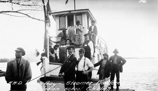 Black and white image of a small two-tiered steamboat moored at a dock with an expanse of water behind. Eleven people are seen standing in the boat, many of them Annishinaabe children. Five men stand beside the boat on the dock in the foreground. Some white text appears at the top and bottom of the image. At top left: May 26th, 1914. At bottom only part of the text is legible: “W.W.D [..] Bay – Steamer Wondere[..]Indian school Children.”