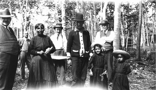 Black and white photograph of, from left to right, foreground: a white man, and Anishinaabe woman, man and two children all slightly spaced apart and facing the camera. Behind them stand three white men at some distance also facing the camera, the one on the right with his arms crossed in front of him, and a stand of trees behind everyone with a structure showing through at the right. In the upper left hand corner is written: June 1, 1914.