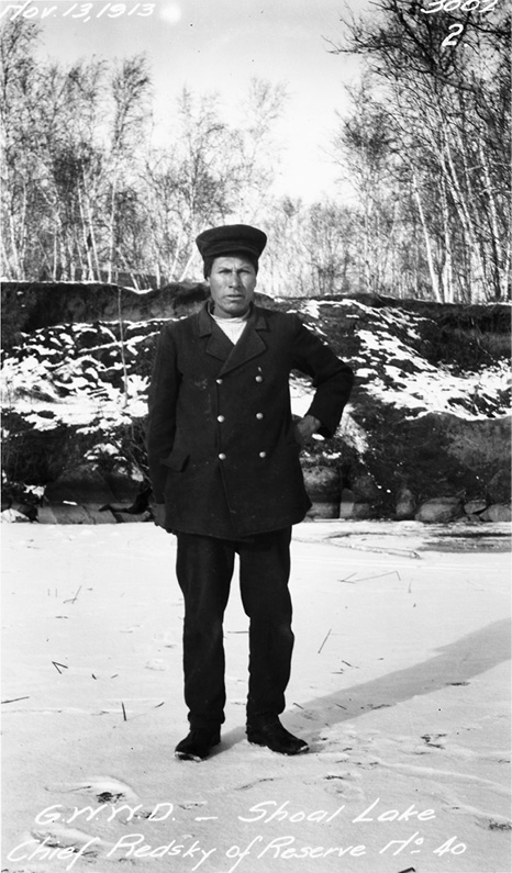 Black and white photograph of Chief Pete Redsky, standing in a Treaty Coat  and hat with one hand on hip. He in a snowy clearing with a partially snow-covered hill and stand of trees behind him. Date in upper left hand corner white script reads: Nov 13, 1913, and at bottom reads: G.W.W.D. – Shoal Lake Chief Redsky of Reserve no 40.