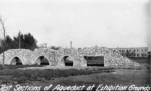 A black and white photograph of a stone wall within which there are four openings, each of a differing shape; from left to right: a peaked arch, a semi-circular arch that meets the ground at its maximum circumference, a circular arch that curves inward to meet the ground just beyond its circumference; and a rectangular-shaped opening with longer width than sides. Behind this structure are some trees at the left, scattered telephone poles, and a low rectangular building behind and to the right. White script at bottom reads: Test Sections of Aqueduct at Exhibition Grounds.