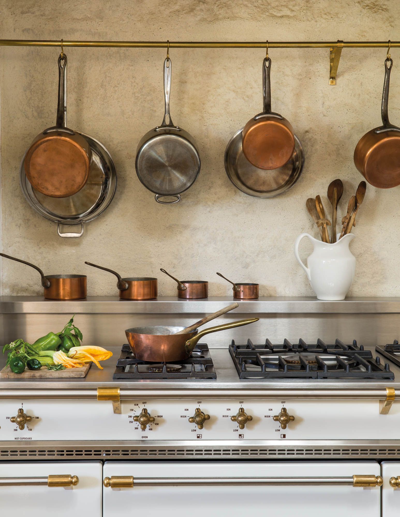 Photo of pots and pans hanging above the range, making them easily accessible for everyday use. Utensils, stored in a white earthenware pitcher are also close at hand.