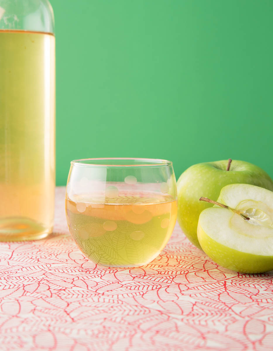 photo of granny smith apple whole and a half, bottle and a glass of apple wine (looks like a white wine) on patterned table cloth and green backdrop