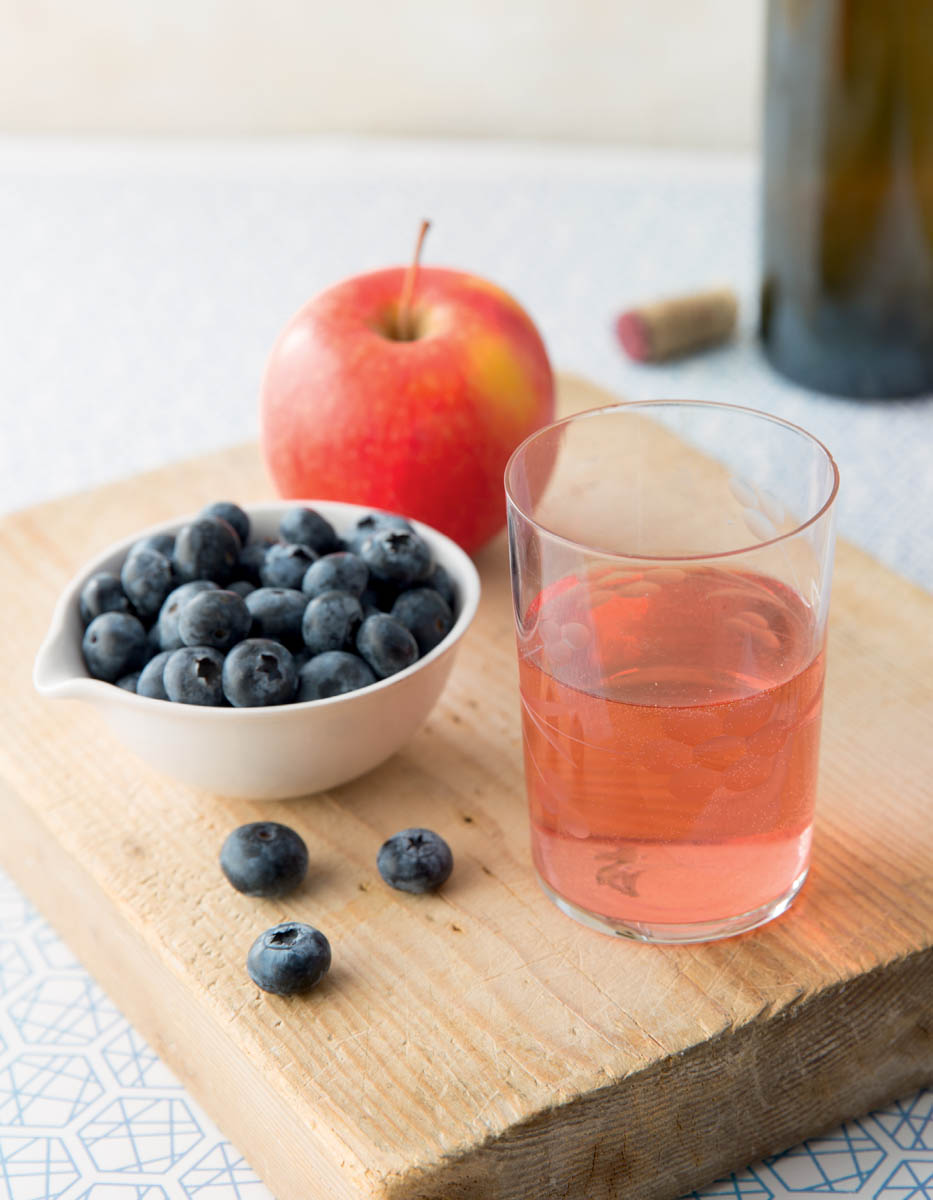 photo of blueberry-apple wine, cup of blueberries, and an apple on a wooden cutting board.