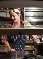 A young woman with her hair swept into a bandana, standing behind a restaurant kitchen counter