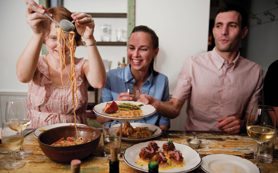 Smiling young people, recognizably some of the staff of Joe Beef, sitting at a large wooden dinner table and sharing family-style meal