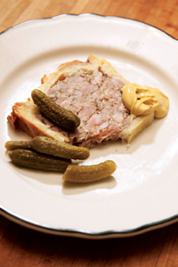 A slice of pate en croute on a plate, accompanied by a handful of small pickles and a large dab of mustard