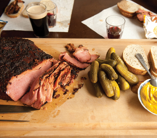 A large wooden board piled with sliced brisket from a huge chunk, a pile of pickles, thick slices of rye bread, and a bowl of yellow mustard. Glasses of red wine and pints of stout beer surround squares of parchment serving as holders for sandwiches made from the spread