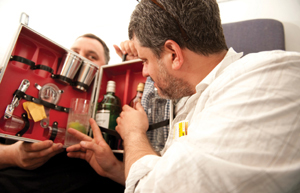 A man turning in a padded seat to pull bottles and glasses from a traveling cocktail case that his companion, in the back seat, is holding through the seats