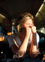 A young woman with her blond hair pulled in a ponytail, her elbows on the table with her hands propped under her chin, seated at a table in a white t-shirt and staring out of the window into the bright morning sun