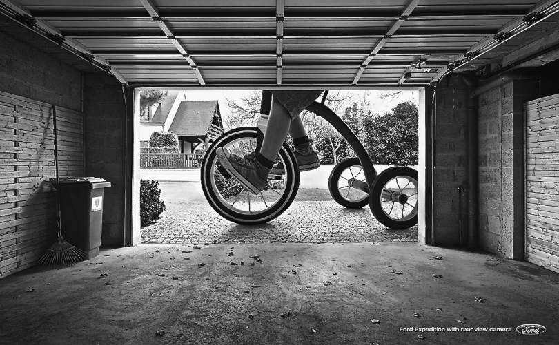 Figure representing an ad campaign for Ford that depicts a magnified view of the lower part of a kid riding a tricycle, as viewed from the garage. The headline reads “Ford expedition with rear view camera.”