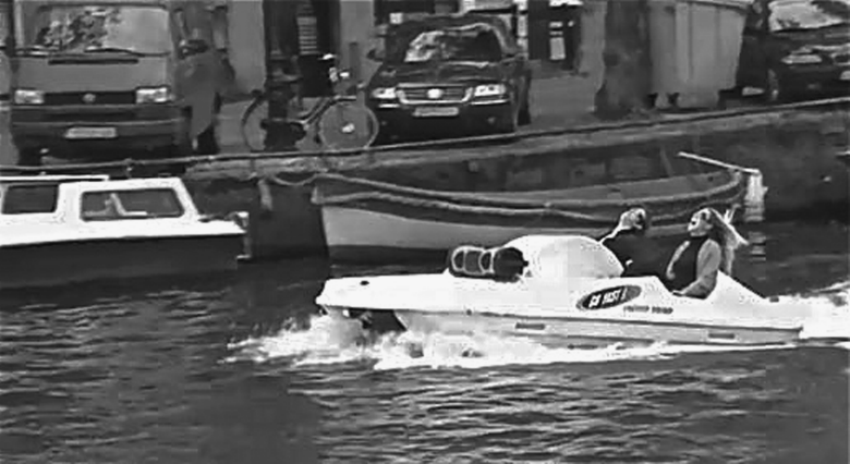 Figure depicting an example of outdoor as an event, where a man and a woman are enjoying on a motorboat in a river. The picture also depicts some cars parked on the road along the river.