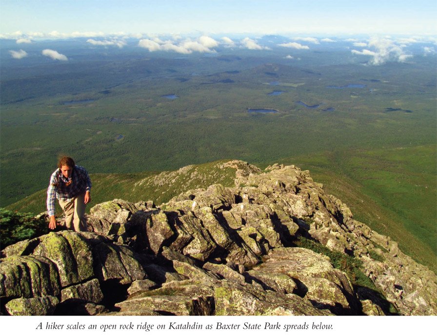 A hiker scales an open rock ridge on Katahdin as Baxter State Park spreads below.