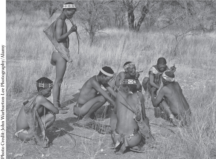 Photograph of a group of seven tribal men in a forest, with six men sitting and smoking together and one man standing near them.