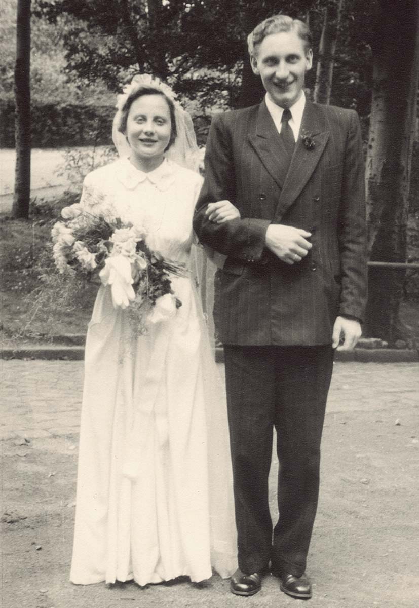 Hochzeitsfoto von Angela Merkels Eltern. Die Mutter (Herlind) trägt ein bis zum Boden reichendes weißes Hochzeitskleid und hält in der rechten Hand einen Blumenstrauß. In der linken ist sie bei dem Bräutigam (Horst Kasner) untergehakt, der einen dunklen Anzug trägt.