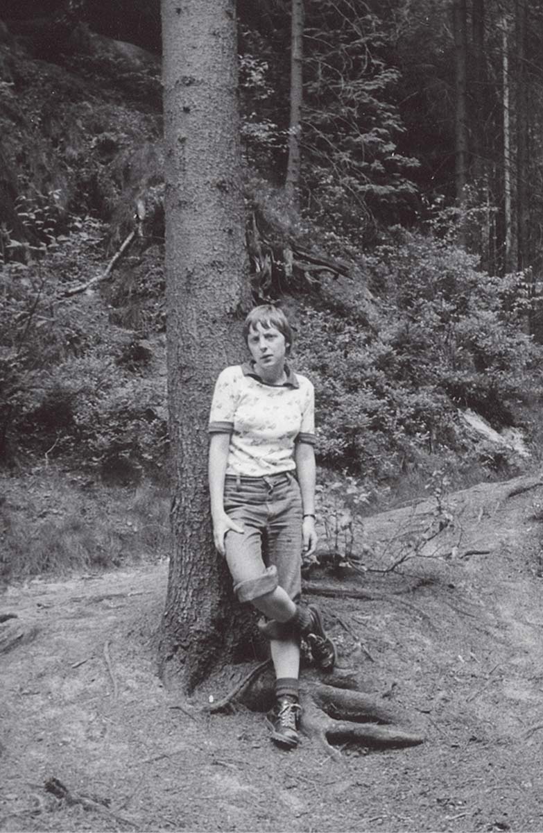 Angela Merkel als Studentin in einem Wald, an einen Baum lehnend, in die Kamera blickend. Sie trägt Wanderschuhe, hochgekrempelte Hosen und eine kurzärmlige Bluse.