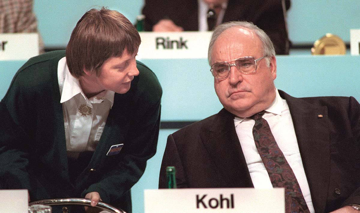 Helmut Kohl auf eine Rednertribüne sitzend, vor ihm sein Namensschild, im Hitnergrund weitere Redner mit Namensschildern, neben ihm, sich zu ihm hinterbeugend Angela Merkel, in weißem Hemd und grüner Jacke