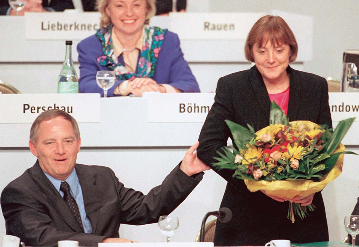 Angela Merkel mit Blumen in der Hand auf einer Rednertribüne, im Hintergrund weitere Redner, jeweils hinter Namensschildern sitzend, neben ihr sitzend Wolfgang Schäuble, sie am Arm halten. 