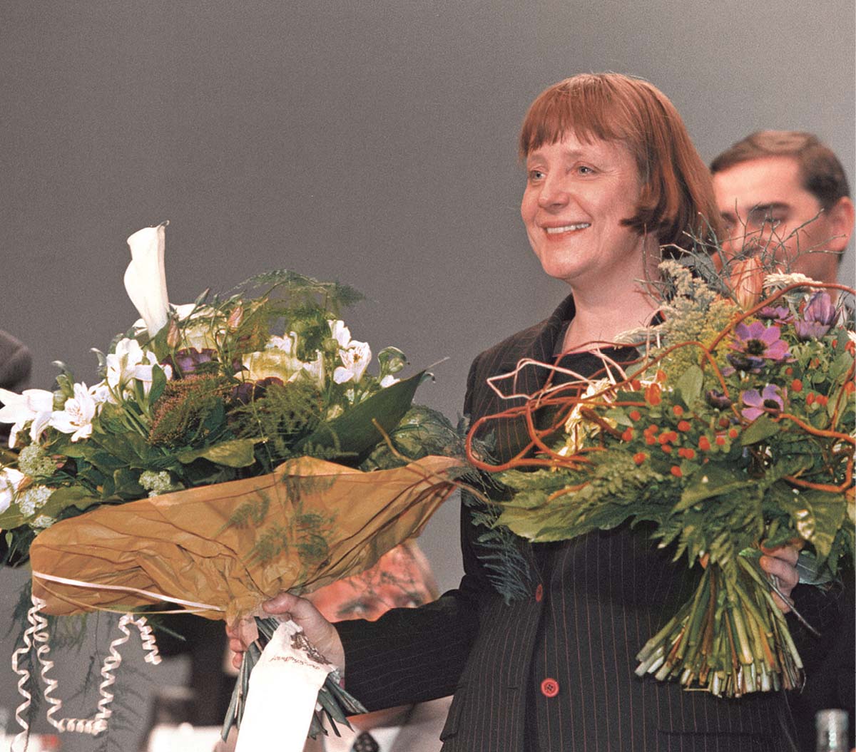 Angela Merkel in dunklem Blazer, glücklich lächelnd, mit zwei Blumensträußen in den Händen