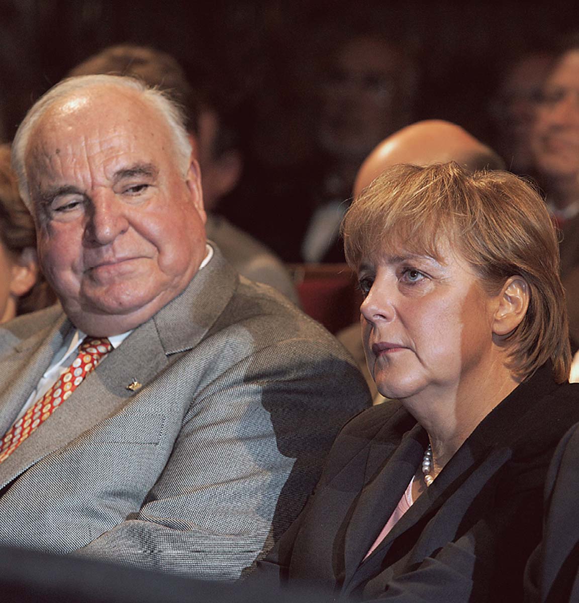 Angela Merkel in dunklem Blazer, nach vorn blickend, neben ihr Helmut Kohl, sie kritisch anschauend