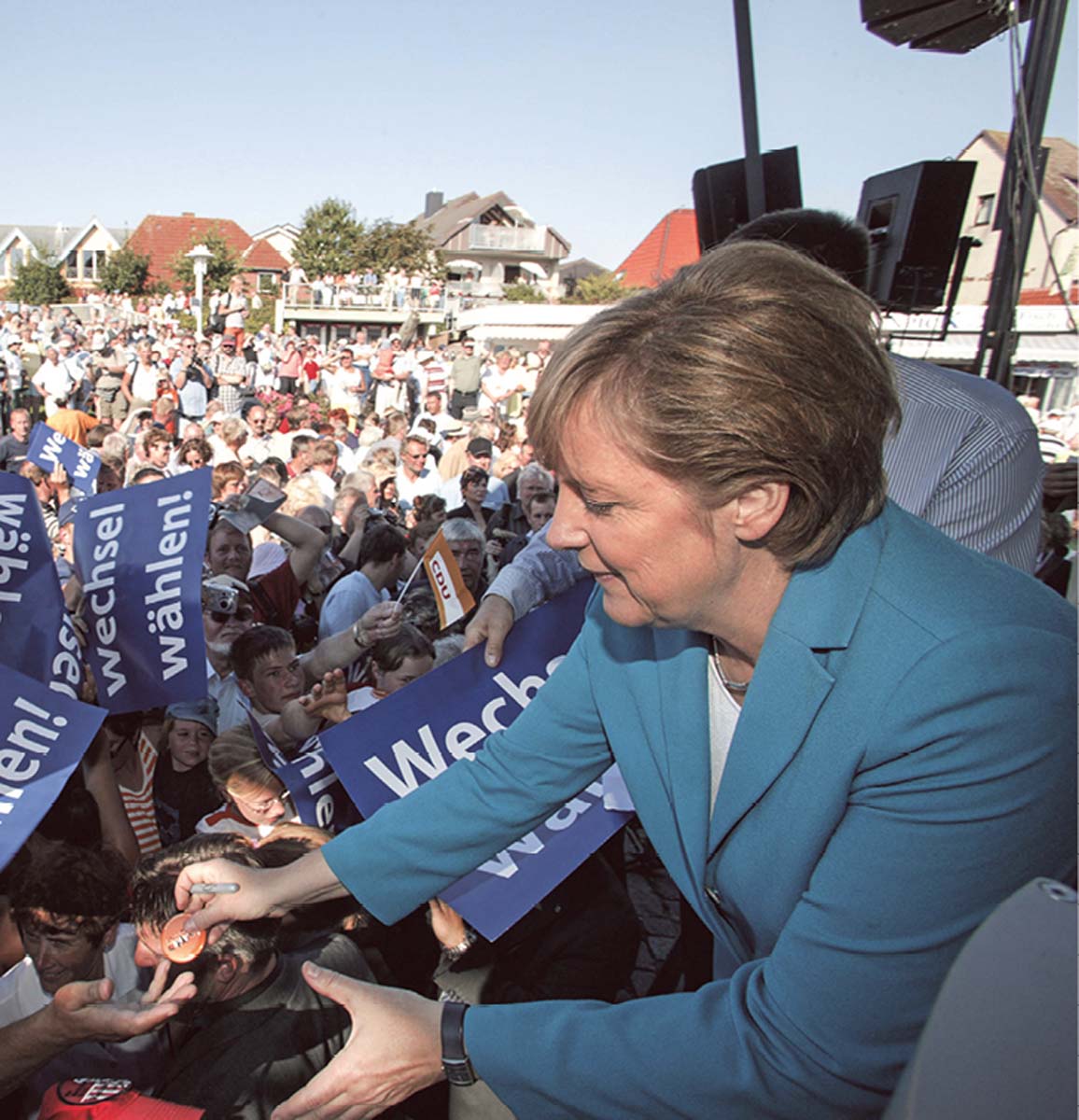 Angela Merkel auf einer Bühne, sich zu einem Zuhörer beugend, vor hunderten Menschen, die  zum Teil Plakate mit der Aufschrift "Wechsel wählen" halten