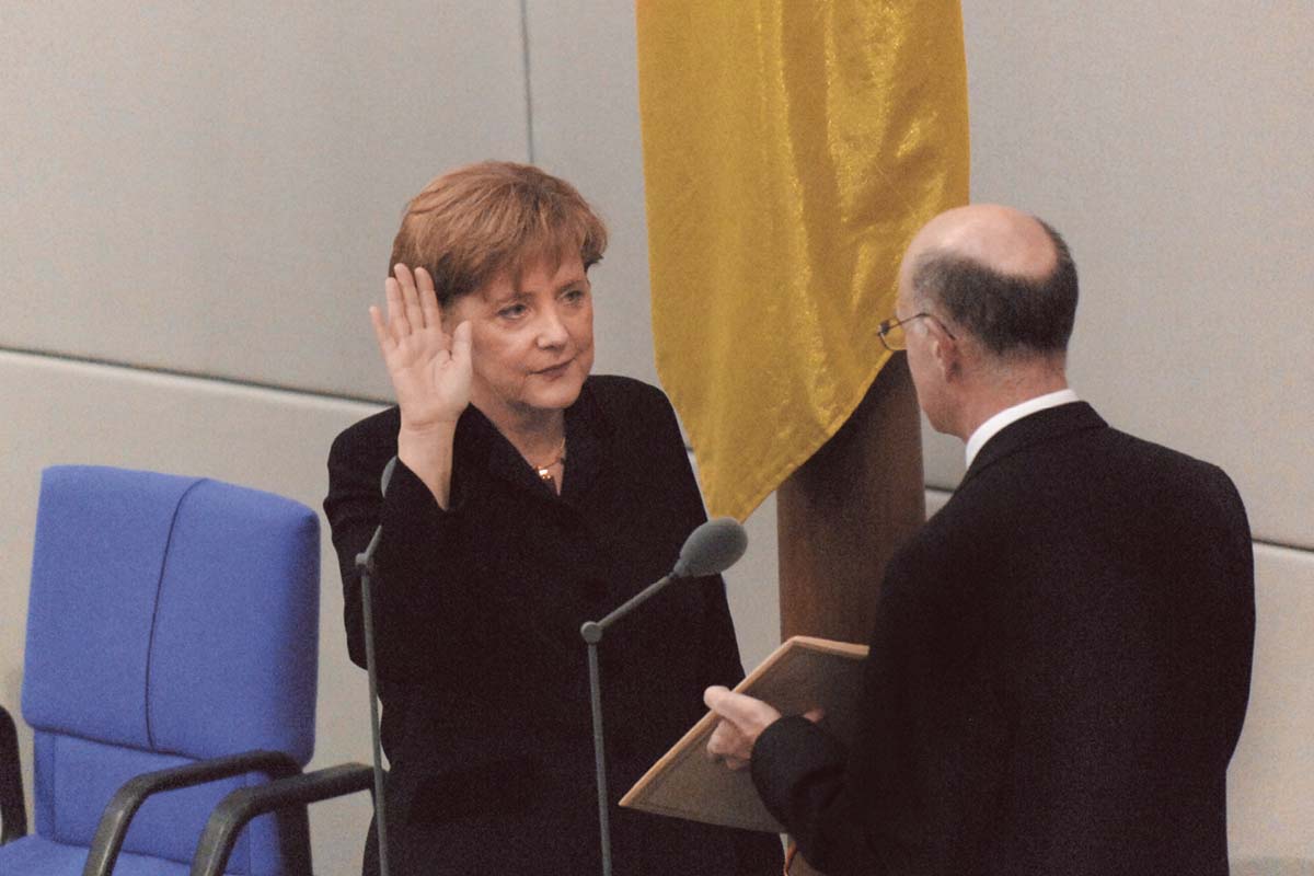 Angela Merkel hebt die rechte Hand, blickt den vor ihr stehenden Mann an, der einen dunklen Anzug trägt. 