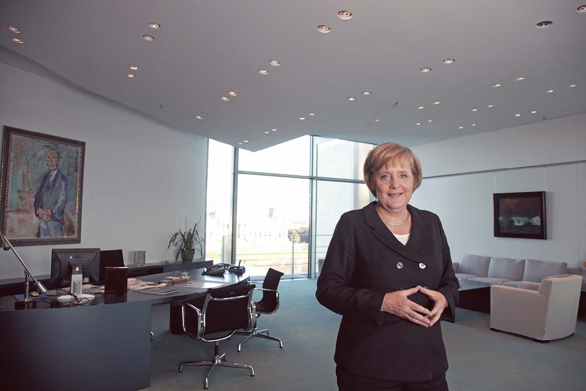 Angela Merkel steht in einem Großen Büro, blickt den Betrachter an, die Hände zu einer Raute verschränkt. Hinter ihrem Schreibtisch hängt ein Bild von Konrad Adenauer