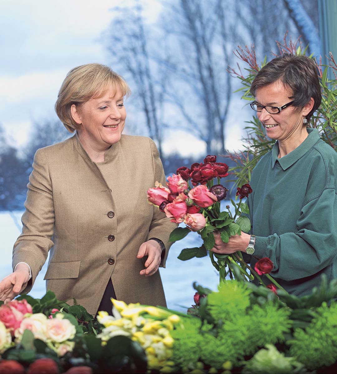 Angela Merkel steht hinter einem Tisch voller Schnittblumen, neben ihr eine Frau, die Rosen zu einem Strauch bindet