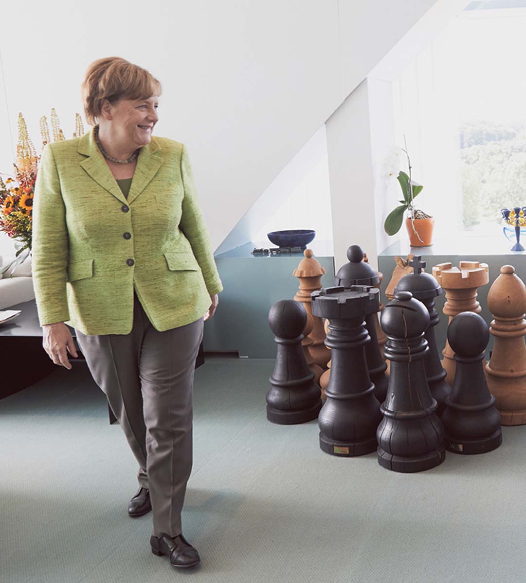 Angela Merkel in einem hellen Raum, neben eihr twa ein Dutzend helle und dunkle hüfthohe Schachfiguren