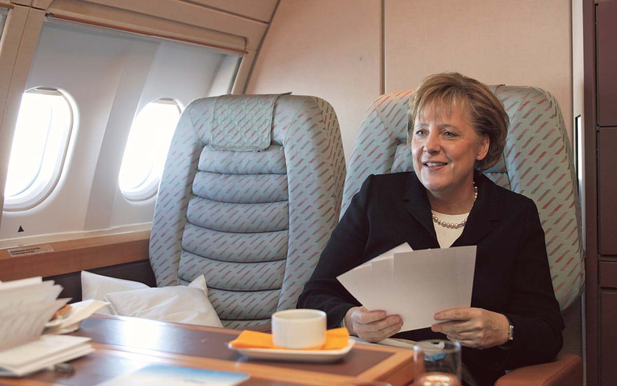 Angela Merkel in einem Flugzeug, an einem Tisch sitzend, der Platz neben ihr ist leer, vor ihr eine Tasse und ein Teller. In den Händen hält sie Papiere.