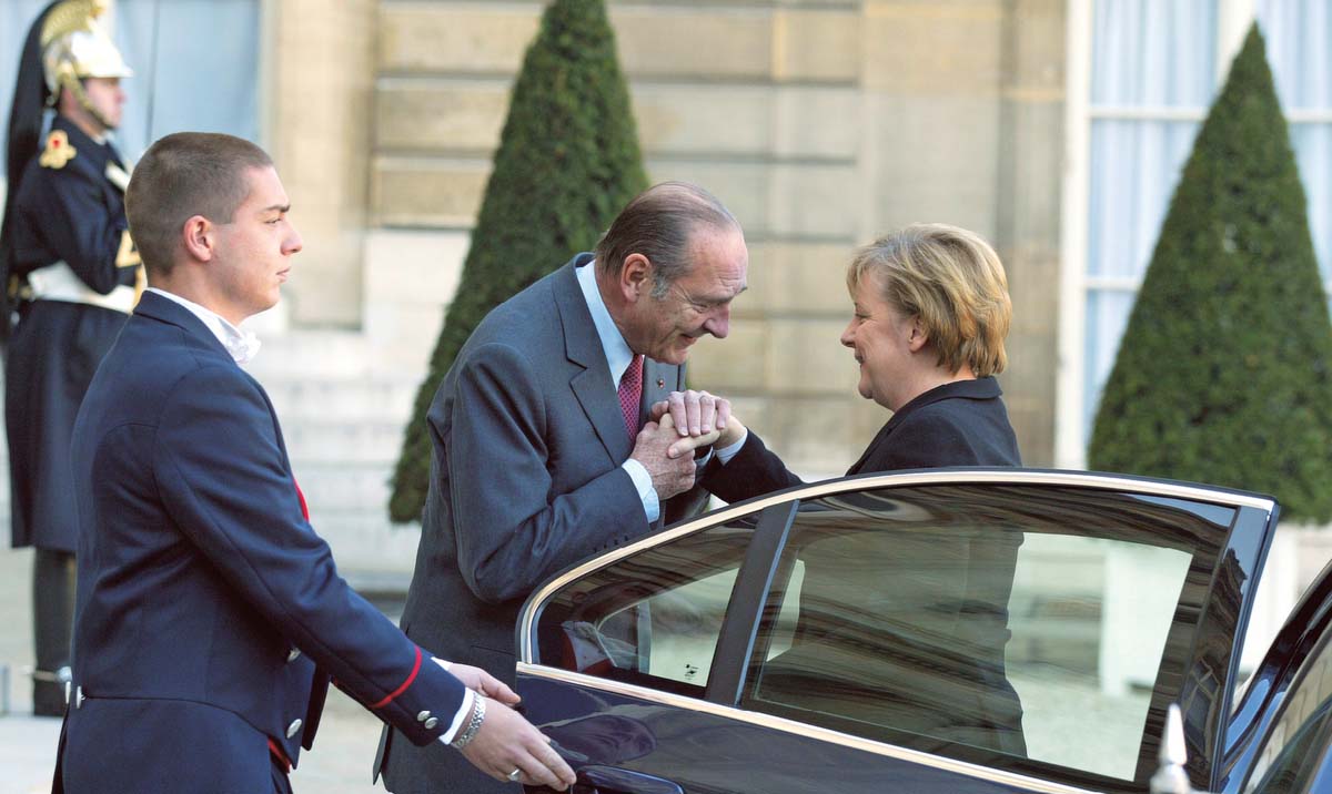 Angela Merkel, lächelnd, soeben aus einem Auto ausgestiegen, ein livrierter Mann hält ihr die Tür auf. Vor ihr steht Jacques Chirac, sich zu ihrer Hand hinterbeugend.