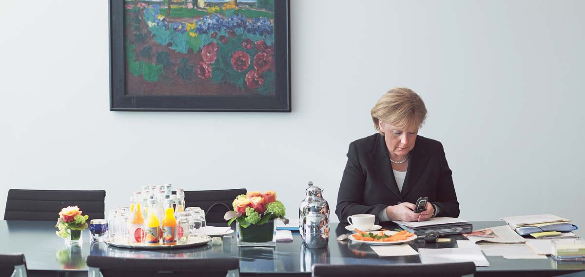 Angela Merkel allein an einem Besprechungstisch, darauf Blumen, Getränke und Papiere. Sie blickt auf ein Handy, das sie in den Händen hält