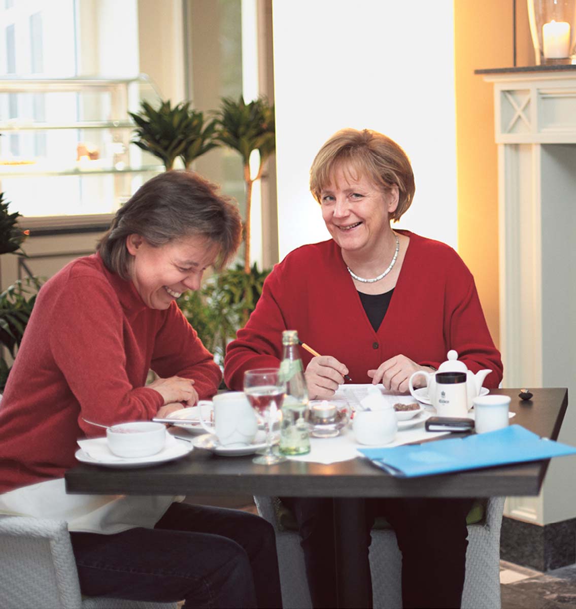 Angela Merkel und Beate Baumann sitzen über Eck an einem quadratischen Tisch, auf dem Gläser, Tassen, Karaffen und Flaschen stehen. Angela Merkel blickt lachend den Betrachter an, Beate Baumann blickt lachend nach unten