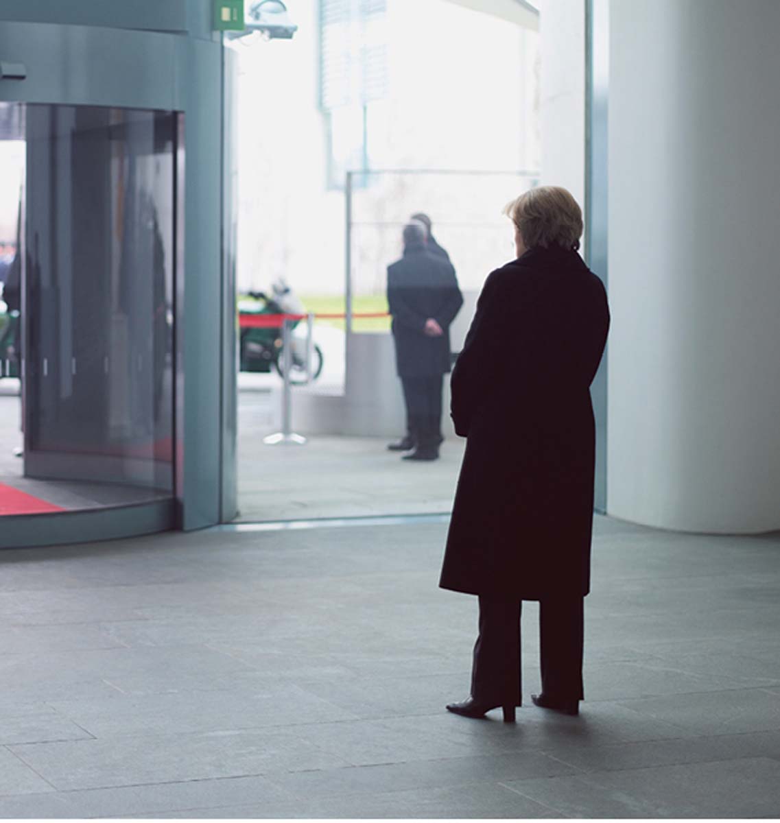 Angela Merkel, von hinten zu sehen, steht allein in dunklem Mantel in einem Foyer und blickt auf die Glastüren vor sich.