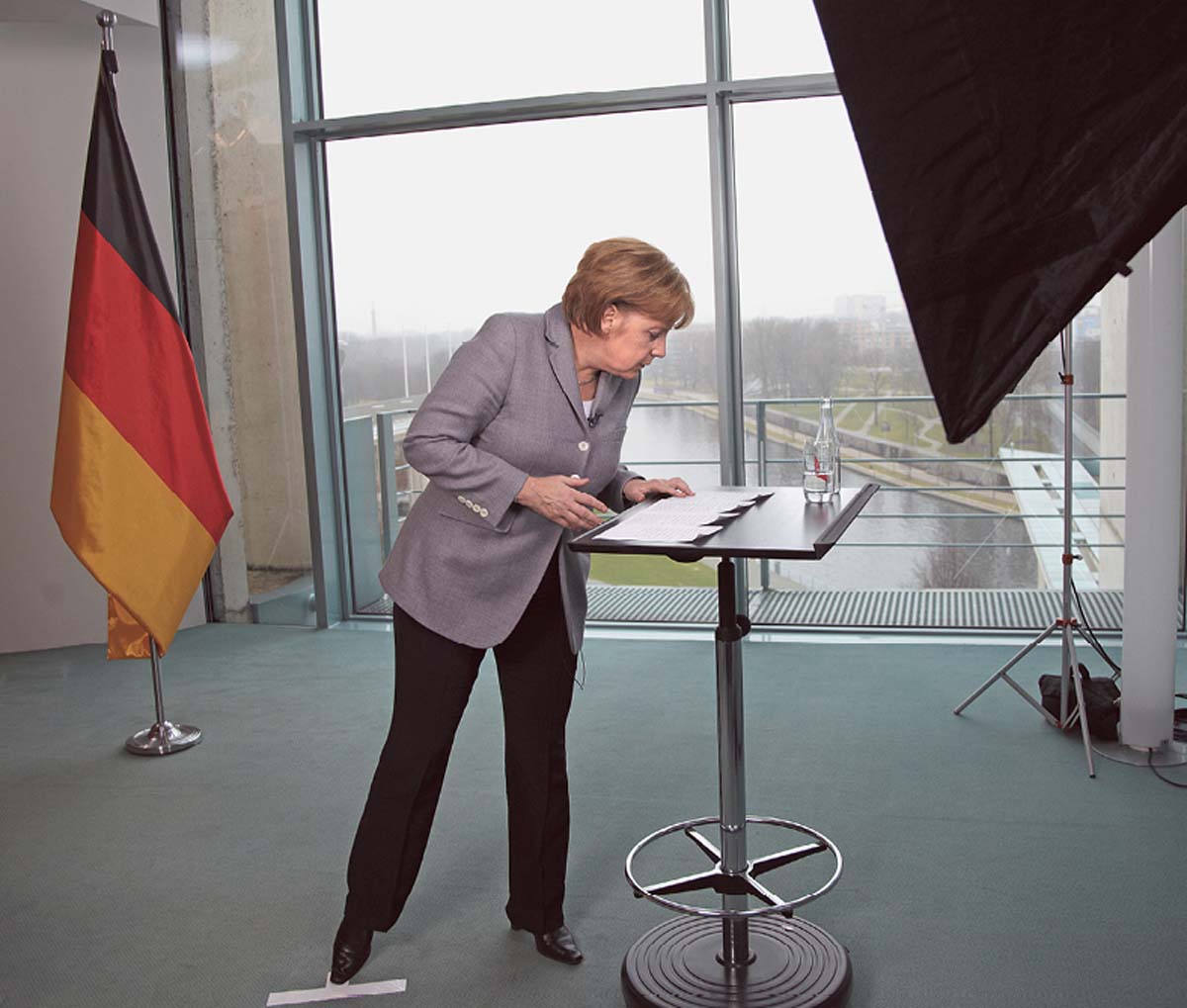 Angela Merkel steht an einem Stehtisch und blickt auf die darauf liegenden Papiere. Im Hintergrund ein bodentiefes Fenster, hinter dem ein Fluss erkennbar ist, und eine Deutschlandfahne